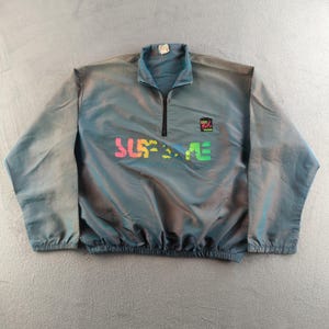 May include: A teal and grey anorak jacket with a half-zip and a stand-up collar. The jacket features the word "SURF" in colourful letters across the front, along with a small logo. The jacket has long sleeves and a drawstring hem.