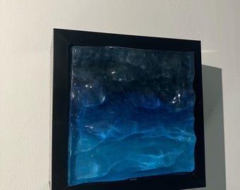 May include: Square artwork featuring a textured, layered design in shades of blue and black, resembling ocean waves. The artwork is encased in a black frame, creating a modern aesthetic. The piece is mounted on a white wall.
