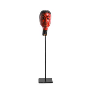 Pode incluir: Uma escultura de cabeça de madeira vermelha e preta em um suporte de metal preto. A cabeça tem um rosto detalhado com traços pintados. A escultura é um item decorativo, provavelmente para decoração de casa ou exibição artística.