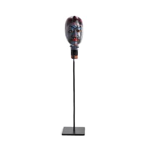 May include: A decorative mask with a blue face, red accents, and red lips is mounted on a black metal stand. The mask has a detailed design and is set against a white background. The stand has a square base.