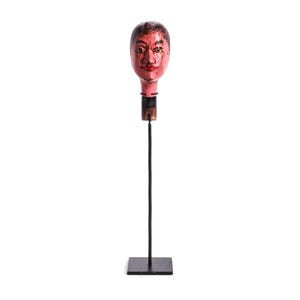 May include: A decorative wooden head sculpture with a painted face, mounted on a black metal stand. The head is painted with red, brown, and black tones. The sculpture has a square base and a long, thin stem.