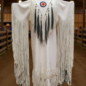 May include: A white dress with long fringed sleeves and hem, displayed on a mannequin. The dress features a decorative neckline with a circular emblem and black fringe accents. The garment is displayed in a rustic wooden structure.