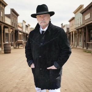 May include: A man wearing a black suede coat and a black hat stands on a dusty street in front of a western town backdrop. The coat has a button closure and pockets. The hat has a wide brim.