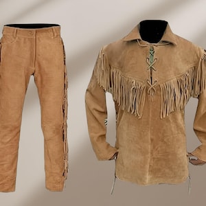 May include: A tan suede leather outfit, including pants and a long-sleeved shirt. The pants have fringe along the sides. The shirt features fringe across the chest and a laced neckline. The outfit is displayed on a neutral background.