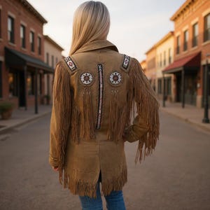 Puede incluir: Chaqueta de ante marrón con flecos y adornos de cuentas. La chaqueta presenta patrones decorativos en los hombros y la espalda, con largos flecos a lo largo de las mangas y el dobladillo. La chaqueta se lleva sobre unos vaqueros azules.