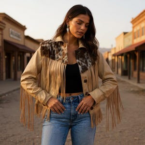 Peut inclure: Veste en cuir beige avec des franges et un col à motifs. La veste a une fermeture boutonnée sur le devant et des manches longues. Le modèle porte un jean bleu et un haut noir.