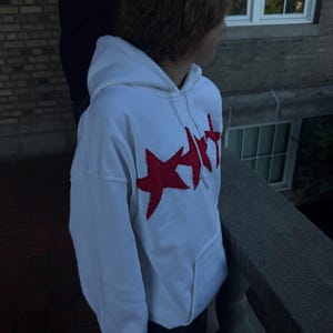 Op de afbeelding: Witte hoodie met een groot rood sterrenontwerp op de voorkant. De hoodie heeft een kangoeroezak en een capuchon met trekkoord. Het ontwerp is een opvallende grafische afbeelding. De hoodie is gemaakt van een zacht, comfortabel materiaal.