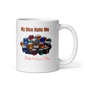 Peut inclure: Mug en céramique blanche avec le texte "My Dice Hate Me" en marron et "But I Love Them" en rouge. La tasse présente une collection de dés colorés en forme de gemmes.