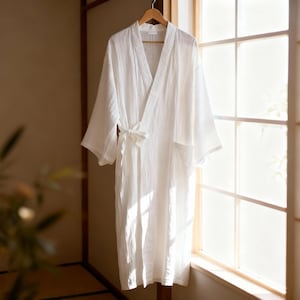 Puede incluir: Una bata blanca y ligera cuelga de una percha de madera cerca de una ventana. La bata tiene un diseño envolvente con cierre de lazo en la cintura y mangas anchas. La tela parece tener textura y la bata está hecha de un material suave.
