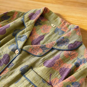 May include: Close-up of a green pajama top with a leaf pattern in shades of purple, orange, and blue. The top has a notched collar with blue trim, a button closure, and a chest pocket. The fabric appears soft and lightweight.