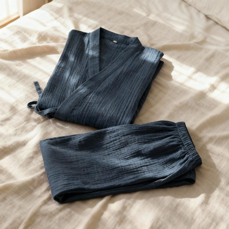 Puede incluir: Un conjunto de dos piezas de ropa de dormir en un tono azul apagado. El conjunto incluye una parte superior tipo envolvente con cierre de lazo y pantalones a juego con cintura el&aacute;stica. La tela parece suave y texturizada, lo que sugiere un ajuste c&oacute;modo y relajado.