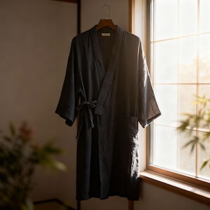 Puede incluir: Una bata gris oscuro con cinturón anudado y bolsillos cuelga de una percha de madera. La bata está hecha de una tela texturizada y tiene un ajuste relajado. La bata está colgada frente a una ventana.