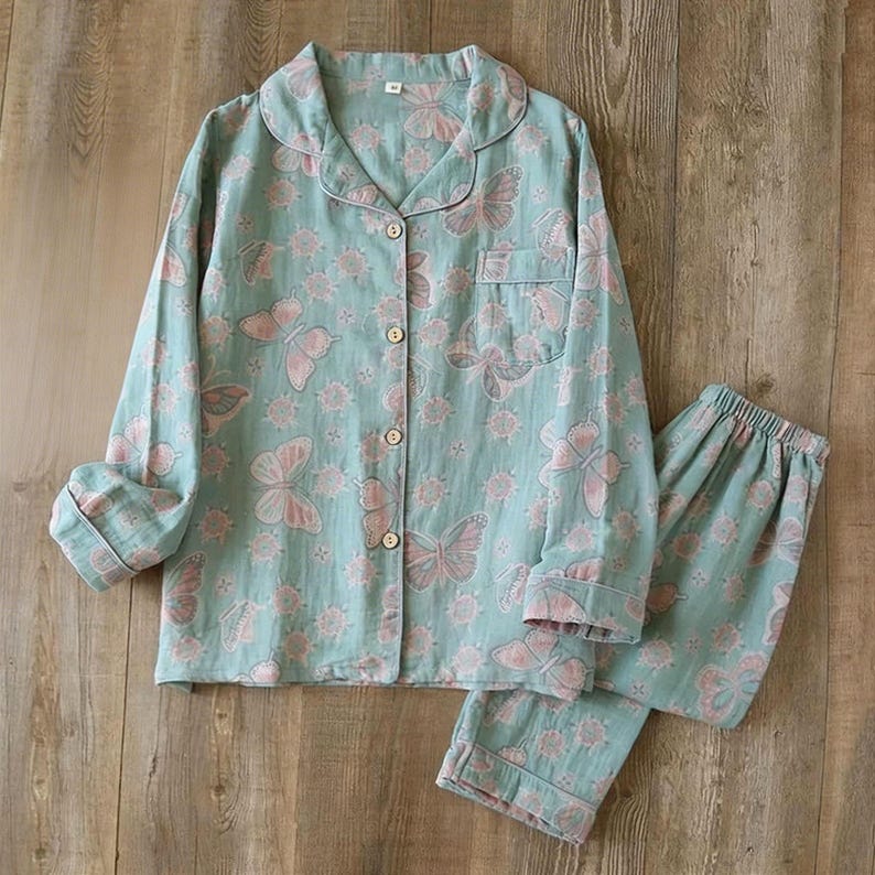 May include: A two-piece pajama set in a light teal color, adorned with a repeating pattern of pink butterflies and floral accents. The long-sleeved button-down shirt has a chest pocket, and the matching pants have an elastic waistband. The set is made of a soft, lightweight fabric.