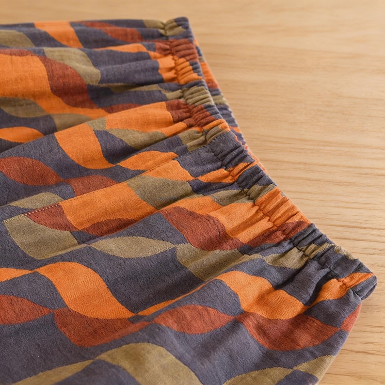 May include: Close-up of patterned fabric with an elastic waistband. The fabric features a repeating wave-like design in shades of orange, brown, and olive green on a dark gray background. The elastic waistband is gathered and visible at the top.