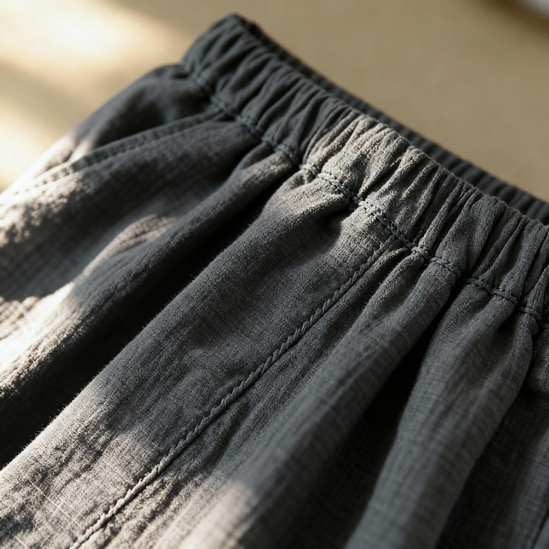 Puede incluir: Primer plano de un pantal&oacute;n de tela gris con cintura el&aacute;stica. La tela tiene un sutil patr&oacute;n texturizado y costuras visibles a lo largo de las costuras. El pantal&oacute;n parece estar hecho de un material suave y ligero.