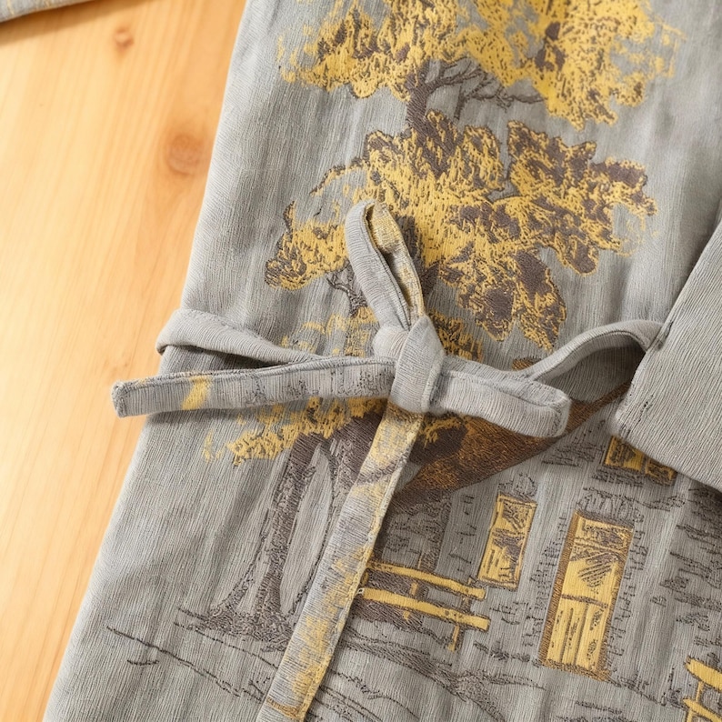 May include: Gray robe with a detailed print of a tree and a building in shades of gold and brown. The robe has a matching tie belt and a textured fabric. The design is reminiscent of traditional Asian art.