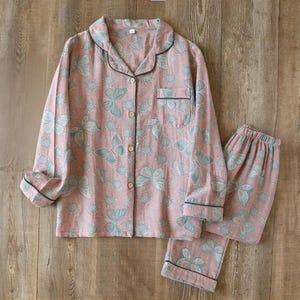 May include: A two-piece pajama set in a soft pink hue, adorned with a repeating pattern of light blue butterflies. The long-sleeved top features a button-down front, a chest pocket, and black trim. The matching pants have an elastic waistband.