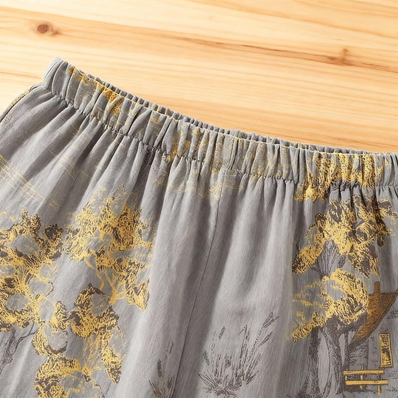 May include: Close-up of gray fabric with an elastic waistband. The fabric features a yellow and gray landscape design with trees and a building. The material appears to be lightweight and suitable for casual wear.