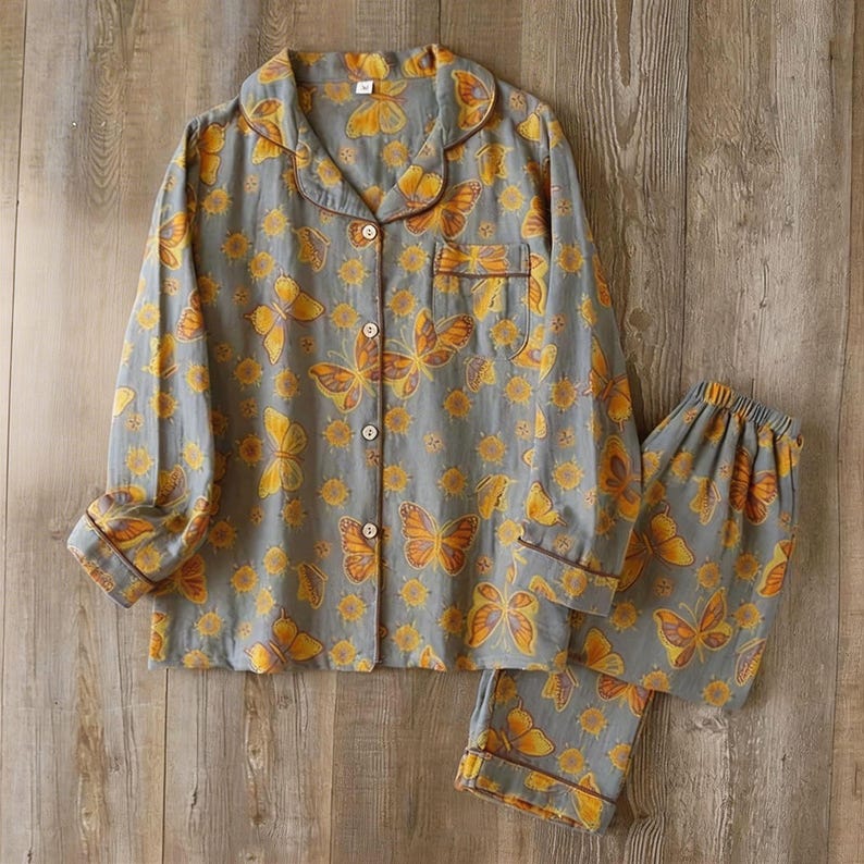 May include: Gray pajama set with a butterfly and floral pattern. The long-sleeved top has a button-down front and a chest pocket. The matching pants have an elastic waistband. The trim is brown.