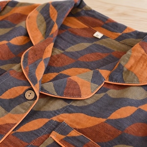 May include: A button-down shirt featuring a repeating wave pattern in shades of orange, dark gray, and olive green. The garment includes a notched collar with orange piping, a single wooden button, and a size M tag at the neckline.
