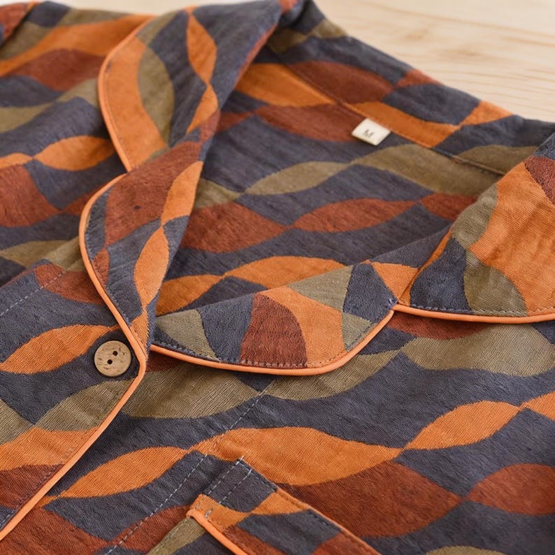 May include: A button-down shirt featuring a repeating wave pattern in shades of orange, dark gray, and olive green. The garment includes a notched collar with orange piping, a single wooden button, and a size M tag at the neckline.