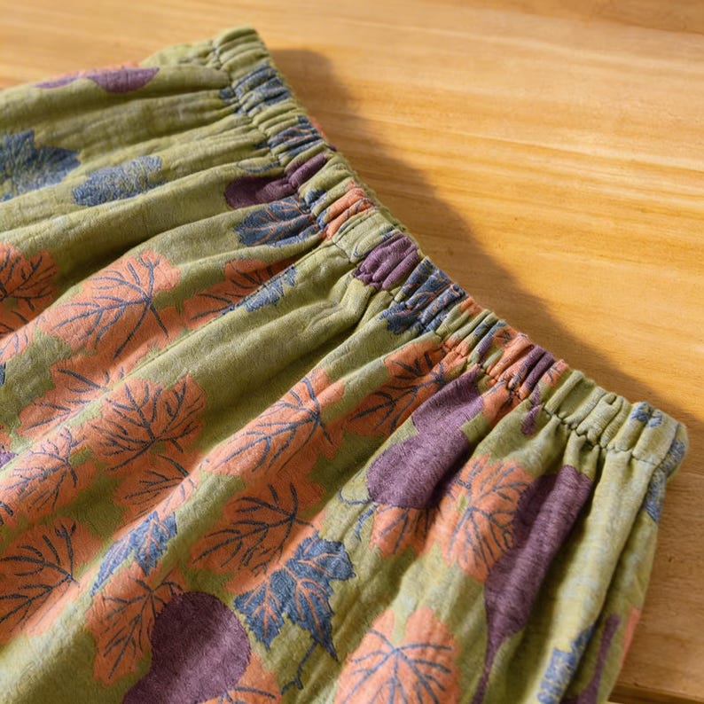 May include: Close-up of olive green fabric with an elastic waistband. The fabric is patterned with large leaf designs in shades of orange, purple, and blue. The leaves are scattered across the material.