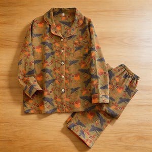 May include: Olive green pajama set with a floral and bird pattern. The long-sleeved top has a button-down front, a chest pocket, and orange trim. The matching pants have an elastic waistband. The fabric features orange and navy blue accents.