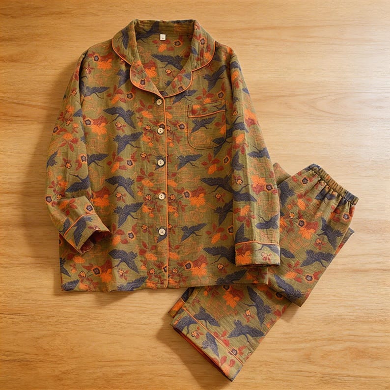 May include: Olive green pajama set with a floral and bird pattern. The long-sleeved top has a button-down front, a chest pocket, and orange trim. The matching pants have an elastic waistband. The fabric features orange and navy blue accents.