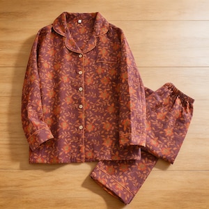May include: A two-piece pajama set in a floral pattern. The set includes a button-down shirt with a chest pocket and pants with an elastic waistband. The fabric is a deep purple with orange and brown floral designs. The shirt has a notched collar and orange trim.