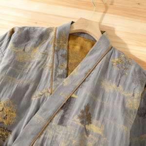 May include: A gray kimono-style robe with a landscape design featuring gold-colored trees and accents. The robe has a contrasting gold-colored interior and brown trim along the edges. The robe is hanging on a wooden hanger.