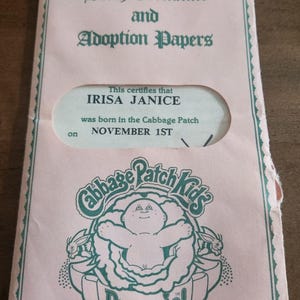 May include: A pink Cabbage Patch Kids birth certificate and adoption papers. The certificate states "IRISA JANICE was born in the Cabbage Patch on NOVEMBER 1ST." The Cabbage Patch Kids logo is at the bottom.