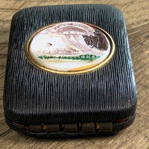 May include: A rectangular black box with a textured surface and a gold-rimmed oval inset. The inset features a miniature landscape scene with a wave, buildings, and a green shoreline. The box has a hinged closure.