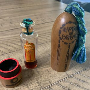May include: A vintage perfume bottle with a colorful label and a wooden container with the text "Maderas de Oriente". The bottle has a decorative top and is filled with a dark liquid. A small wooden container with a red rim is also present.