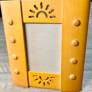 May include: A light brown wooden picture frame with a rectangular center opening. The frame features a cutout sun motif at the top and bottom edges, and four small circular wooden knobs arranged vertically along each side of the frame.