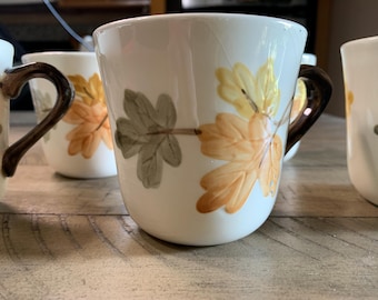 May include: White ceramic mugs with a fall leaf design in shades of yellow, orange, and green. The mugs have brown handles and are set on a wooden surface. The mugs are part of a set.