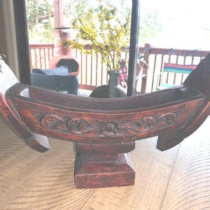May include: A dark brown, wooden decorative boat-shaped object with intricate carvings. The boat rests on a square base and features detailed floral and scrollwork designs. The ends of the boat are curved upwards, adding to its unique shape.