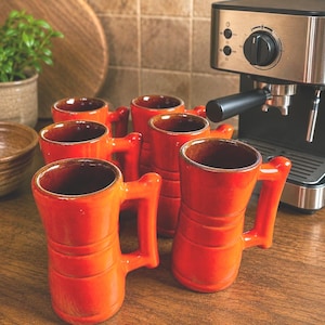 May include: A set of five bright orange ceramic mugs with a glossy finish, each featuring a cylindrical body with horizontal ridges and a sturdy rectangular handle. The mugs are arranged in a cluster, showcasing their vibrant color and smooth texture.