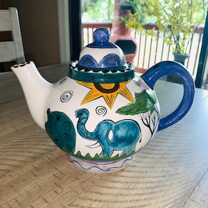 May include: A hand-painted ceramic teapot with a blue handle and lid. The white body features a whimsical design of a blue elephant, sun, and trees. The teapot has a blue spout and a decorative lid with a blue knob.