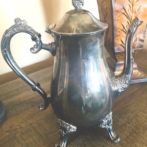 May include: A silver-colored teapot with ornate detailing. The teapot has a curved handle, a spout, and a lid with a decorative finial. It stands on three decorative feet. The teapot is likely vintage or antique.