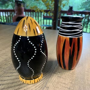 May include: Two decorative ceramic vases. One is egg-shaped, black with white dots and yellow accents. The other is cylindrical, with orange and black vertical stripes and white horizontal lines.