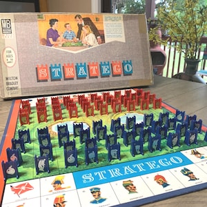 May include: Vintage Stratego board game by Milton Bradley. The game board features red and blue pieces arranged on a green field. The box shows a family playing the game. The game is for ages 8 to adult.