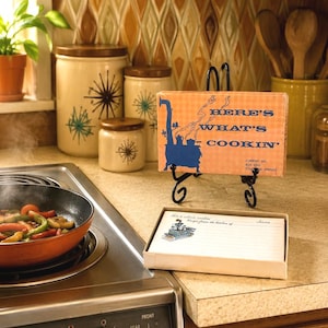 May include: A vintage recipe card box with an orange checkered pattern and a blue illustration of a wood-burning stove. The box features the text Here's What's Cookin' and Current Inc. Box 2243 Colorado Springs. A matching recipe card is displayed in front.