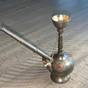 May include: An antique metal hookah with a brass bowl and accents. The hookah has a long, angled stem and a round base. Decorative red stones are set into the body of the hookah.