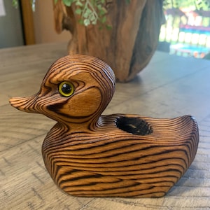 May include: A wooden duck figurine with a detailed wood grain pattern. The duck has a yellow eye and a small, dark hole on its back. The figurine is a decorative item, likely for home decor.