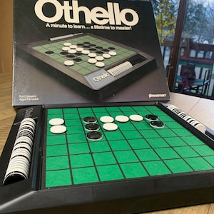 May include: Othello board game with a black frame and a green playing surface. The game includes black and white circular game pieces. The box reads "Othello, A minute to learn... a lifetime to master!" and is black with white lettering.