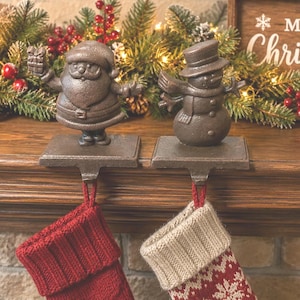 May include: Two dark brown cast iron stocking holders featuring a Santa Claus figure holding a gift and a snowman figure wearing a top hat and scarf. Each figure stands on a flat rectangular base with a hook extending downward for hanging.