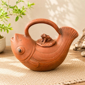 May include: A terracotta teapot shaped like a fish with a curved handle forming the top fin. The lid features a small sculpted frog sitting on a lily pad. The body of the fish has etched scale details and a prominent eye, with a spout at the mouth.