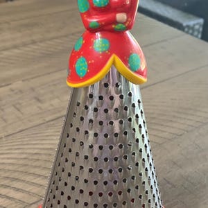 May include: A novelty kitchen grater shaped like a woman in a red dress with green polka dots. The grater has a stainless steel cone with numerous holes and a red base. The woman's head is light-colored with brown hair.