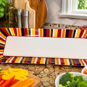 May include: A rectangular serving platter with a solid white center and a wide border featuring vertical stripes in vibrant shades of red, orange, yellow, green, blue, and purple. The platter has a glossy finish and rounded corners.