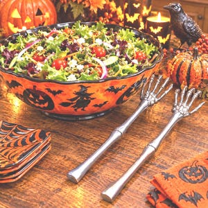 May include: A large orange ceramic bowl with black silhouettes of pumpkins, witches, and bats, filled with a green salad. Two silver-toned metal salad servers shaped like skeletal hands rest beside the bowl, accompanied by small square plates.